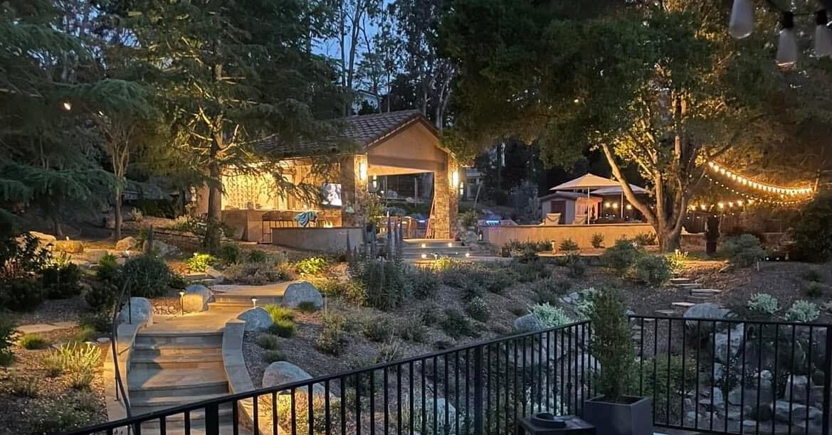 Backyard pavilion, uplit trees, and lit garden steps at dusk