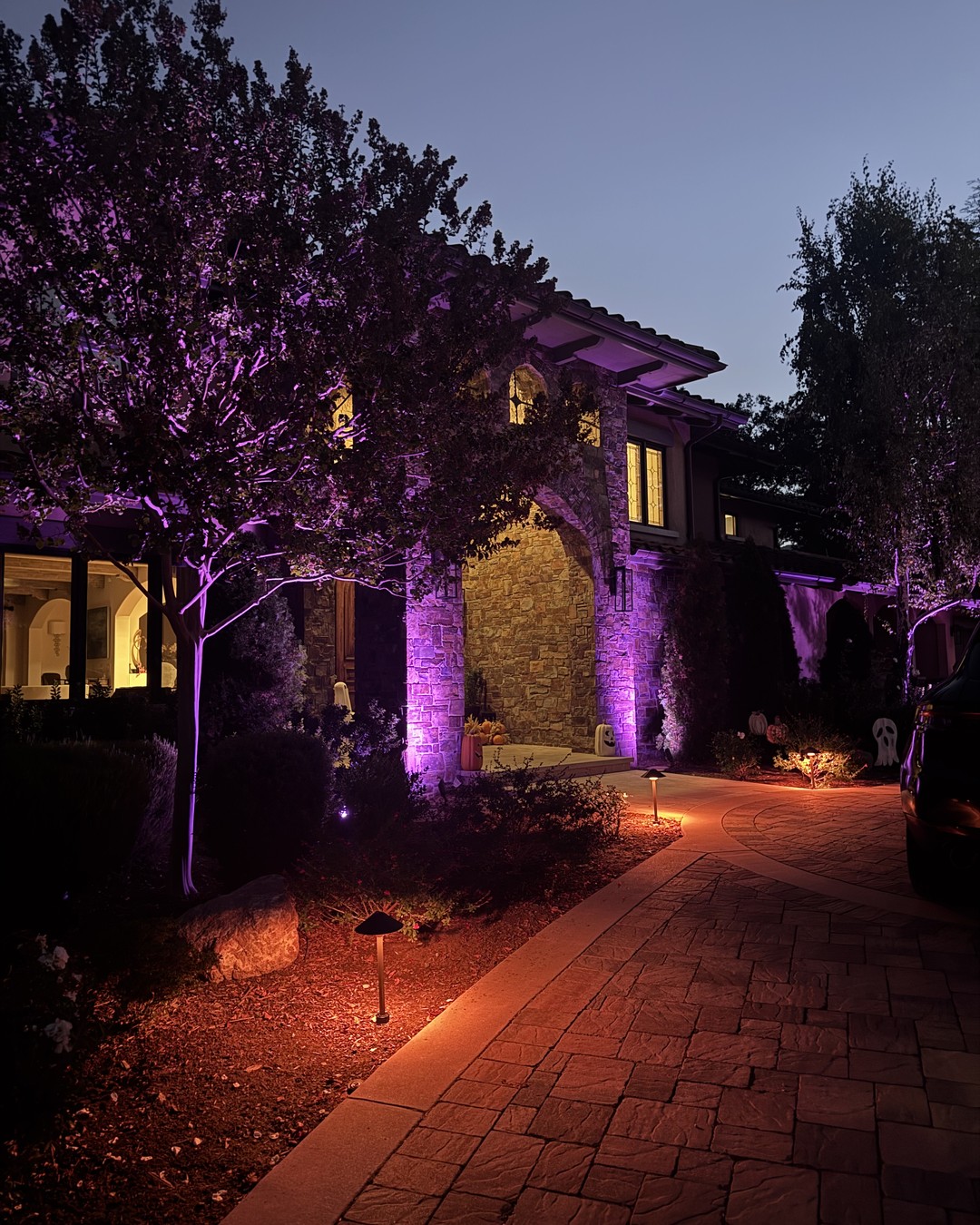 Stone home entry at dusk lit in purple, with warm amber path lights along a paver driveway and small ghost decorations by the door